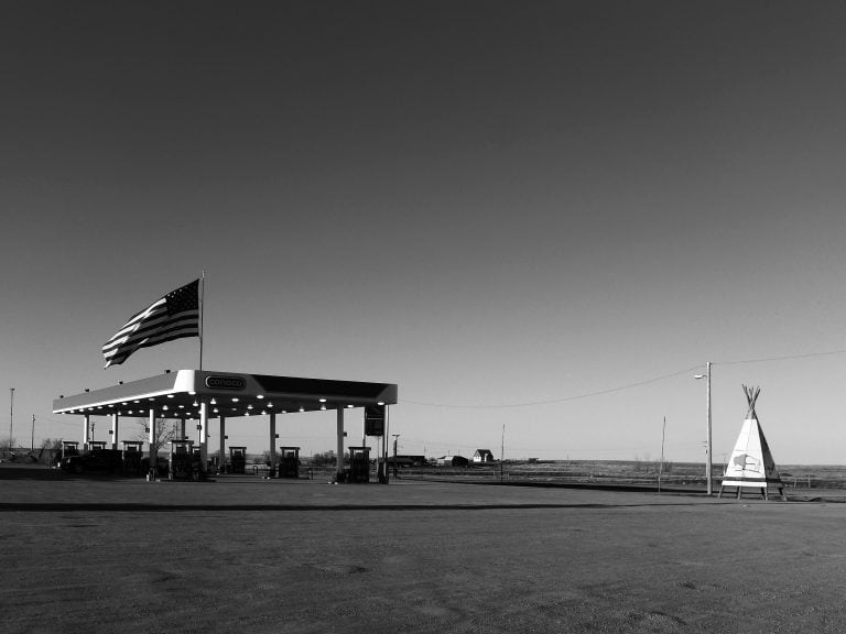 Black and white photo of Conoco gas station with large American flag. Nearby is an imitation of a teepee.